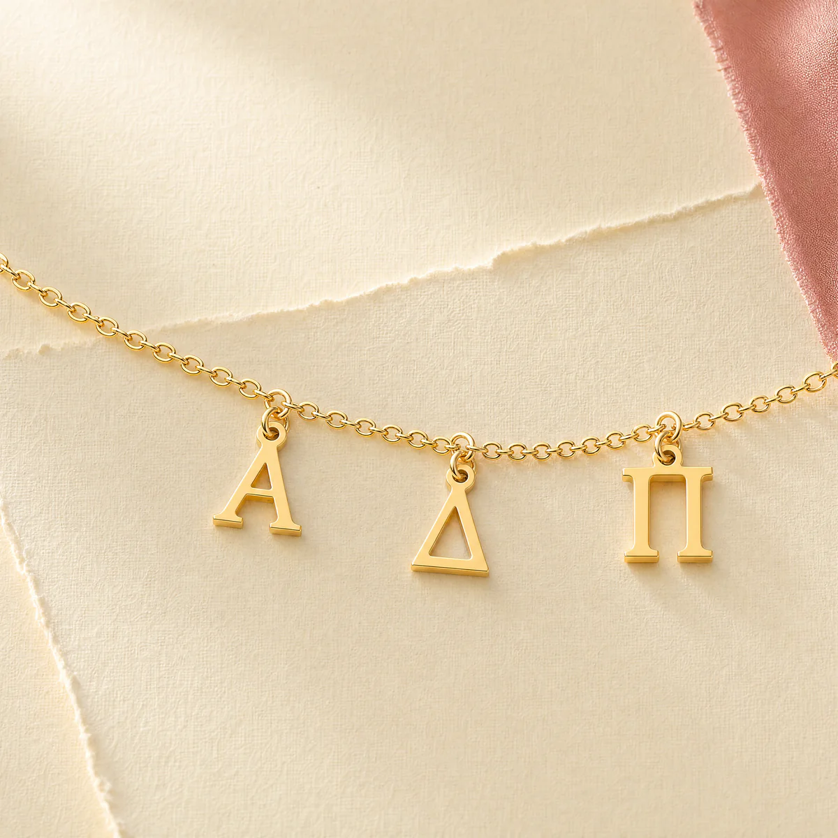 Alpha Delta Pi gold letter pendants arranged across cream paper and blush fabric